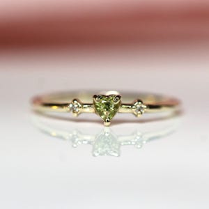 May include: A delicate gold ring featuring a heart-shaped, light green gemstone at the center. Two small, clear gemstones flank the heart. The ring is set against a soft pink background, with its reflection visible below.