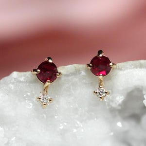 May include: A pair of gold stud earrings, each featuring a round, deep red gemstone set in a four-prong mount, with a smaller, clear, round-cut diamond-like stone suspended directly below the larger red stone on a thin gold bar.