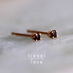 May include: A pair of gold stud earrings featuring small, round, dark black stones set in four-prong settings. The text "liesel love" with a small arrow graphic is positioned below the earrings. The earrings have thin, straight posts.