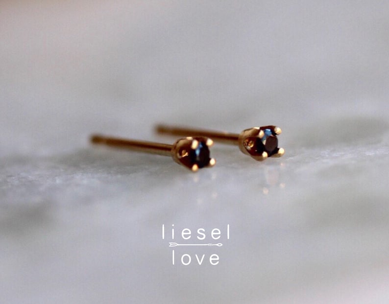 May include: A pair of gold stud earrings featuring small, round, dark black stones set in four-prong settings. The text "liesel love" with a small arrow graphic is positioned below the earrings. The earrings have thin, straight posts.