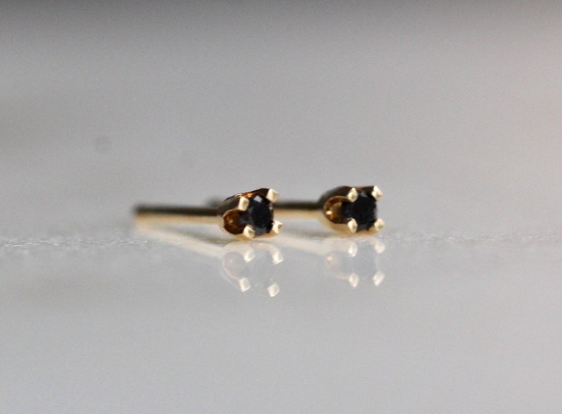 May include: A pair of stud earrings featuring small, round, black stones set in four-prong gold metal settings. The earrings have straight posts extending from the back of the settings, designed for pierced ears, and are shown in a side profile view.