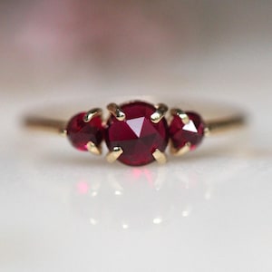 May include: A gold ring featuring three round, faceted deep red rubies set in a horizontal row. The center ruby is larger than the two flanking stones, and each is secured by four small gold prongs on a thin, polished gold band.