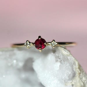May include: A gold ring featuring a central round-cut deep red ruby gemstone held in a four-prong setting, flanked by two smaller clear white diamonds, each set in a four-prong mount on a thin, polished gold band.