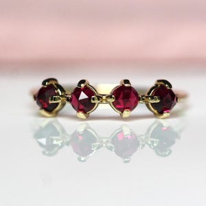 Puede incluir: Un anillo de oro con cuatro piedras preciosas redondas, facetadas y de color rojo intenso dispuestas en una fila horizontal. Cada piedra está asegurada por cuatro pequeñas garras, creando una línea de color continua en el anillo.