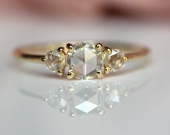 May include: A gold ring featuring a central round rose-cut diamond flanked by two smaller round rose-cut diamonds. The stones are set in yellow gold prongs on a thin, polished yellow gold band. The ring is captured with a clear reflection below.