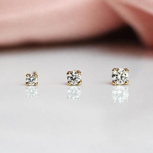 May include: Three round-cut clear stud earrings set in gold prongs, arranged in a horizontal row from smallest to largest. Each earring features a single faceted stone that reflects light, creating a brilliant sparkle against the smooth surface.