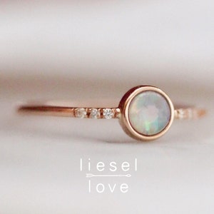 Puede incluir: Un delicado anillo de oro rosa con una piedra de ópalo blanco y una fila de pequeños diamantes.
