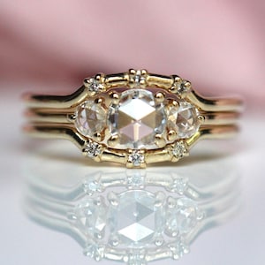 Puede incluir: Un anillo de oro con una gran piedra central redonda facetada flanqueada por dos piedras redondas más pequeñas. La banda está decorada con seis piedras redondas diminutas engastadas en un marco decorativo que rodea las tres piedras.