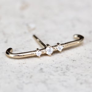 Puede incluir: Un pendiente de aro de oro con tres pequeños diamantes en fila. El aro es curvo y tiene un pequeño poste para la perforación.
