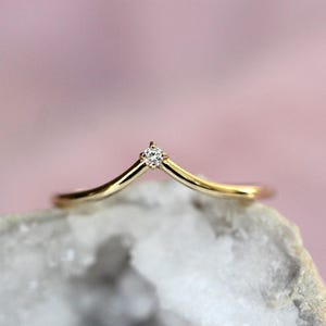May include: A gold V-shaped ring featuring a single small round clear gemstone set at the center point of the band. The band has a smooth, polished finish and follows a chevron silhouette, tapering to a point where the stone is securely mounted.