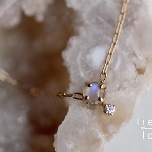 May include: A delicate gold chain necklace featuring a moonstone pendant and a small diamond accent. The necklace is displayed on a white, textured surface.