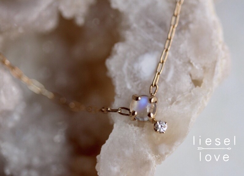 May include: A delicate gold chain necklace featuring a moonstone pendant and a small diamond accent. The necklace is displayed on a white, textured surface.