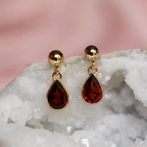 May include: A pair of gold stud earrings featuring dangling teardrop-shaped deep red gemstones. Each earring consists of a smooth gold ball post connected to a faceted red stone set in a gold frame, creating a classic drop design.