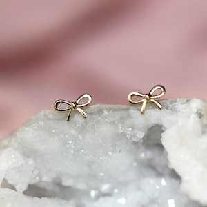 May include: A pair of gold bow-shaped stud earrings on a white rock surface.