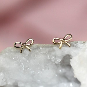 May include: A pair of gold-toned stud earrings shaped like small bows. Each earring features a delicate, open-loop design with two short tails extending downward from the center knot, creating a symmetrical and polished appearance for the jewelry.