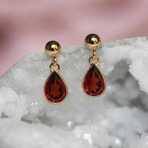 May include: A pair of gold stud earrings featuring dangling teardrop-shaped red garnet gemstones. Each earring consists of a polished gold sphere stud connected to a faceted, deep red pear-cut stone set in a thin gold frame, creating a drop design.