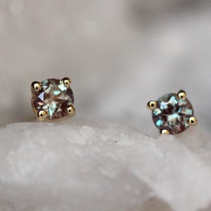 May include: A pair of stud earrings featuring round, faceted alexandrite gemstones set in four-prong yellow gold settings. The stones exhibit a color-changing appearance with visible hues of teal, blue, and purple, securely held by gold prongs.