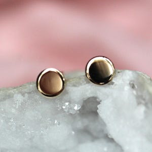 May include: A pair of circular stud earrings with a polished gold-tone finish. The earrings feature a smooth, reflective surface and are positioned on a rough, white crystalline mineral base with a textured, jagged appearance and translucent edges.