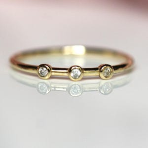May include: A thin gold band ring featuring three small, round, clear stones set in individual bezel settings along the top. The metal has a polished, reflective finish, and the stones are spaced evenly across the front of the ring band.