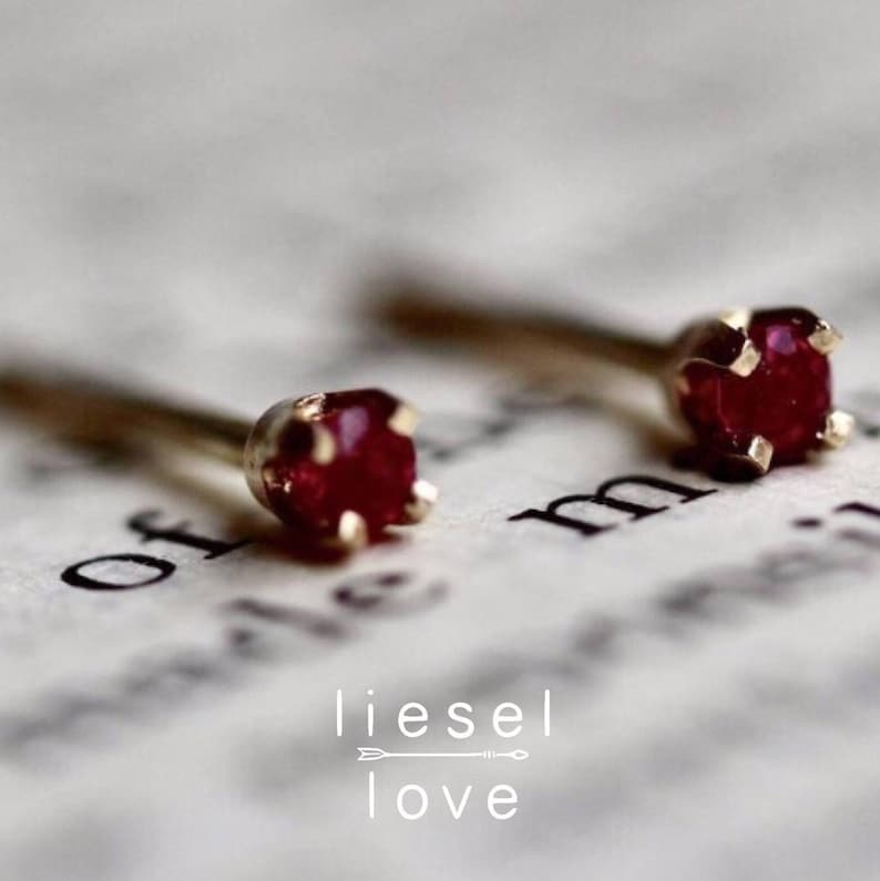 May include: A pair of small gold stud earrings with a single red gemstone in each. The earrings are on a white surface with the text "of" and "m" visible.