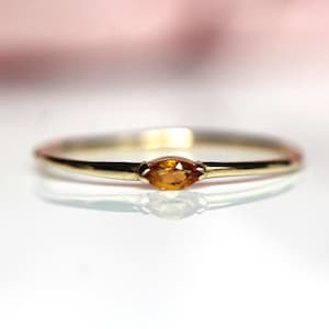 May include: A thin gold band ring featuring a single marquise-cut amber-colored gemstone set horizontally in the center. The metal has a polished finish, and the gemstone displays a warm, translucent orange-yellow hue with faceted surfaces.