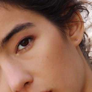 May include: A small, delicate stud earring featuring a tiny, clear, round-cut gemstone set in a simple metal post. The earring is worn in the earlobe, appearing as a subtle, minimalist point of light against the skin, providing a refined accent.