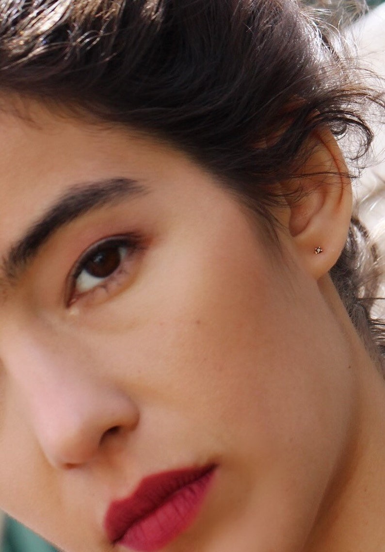 May include: A small, delicate stud earring featuring a tiny, clear, round-cut gemstone set in a simple metal post. The earring is worn in the earlobe, appearing as a subtle, minimalist point of light against the skin, providing a refined accent.