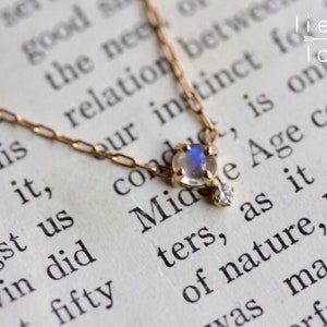 May include: A delicate gold chain necklace with a moonstone pendant and a small diamond accent. The necklace is lying on a book with text that reads "Midge Age could only be as it could of nature, because it was made fully".