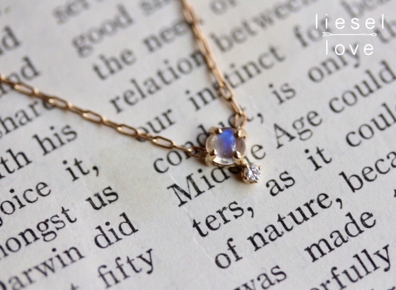 May include: A delicate gold chain necklace with a moonstone pendant and a small diamond accent. The necklace is lying on a book with text that reads "Midge Age could only be as it could of nature, because it was made fully".