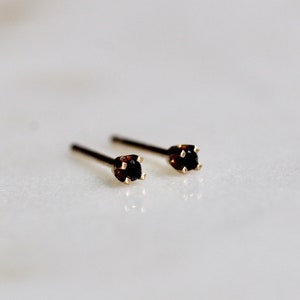 May include: A pair of stud earrings featuring small, round, dark black gemstones set in four-prong gold metal settings. Each earring has a thin, straight post extending from the back of the setting, designed for insertion into a pierced earlobe.