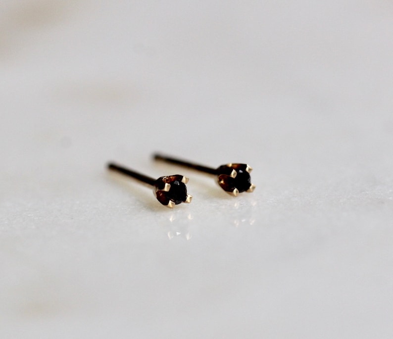 May include: A pair of stud earrings featuring small, round, dark black gemstones set in four-prong gold metal settings. Each earring has a thin, straight post extending from the back of the setting, designed for insertion into a pierced earlobe.