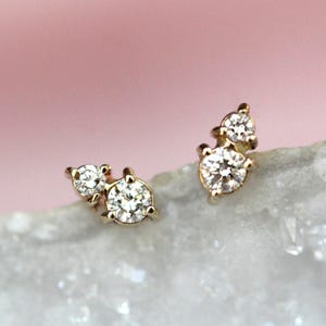 May include: A pair of gold stud earrings, each featuring two round clear stones set in a vertical cluster. The stones are held in place by small gold prongs, creating a delicate and reflective design that catches the light with a brilliant sparkle.