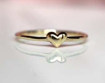 May include: A thin, polished gold band ring featuring a small, centered, three-dimensional heart charm. The metal surface is smooth and reflective, catching the light to create a bright, metallic sheen across the heart and the circular band.