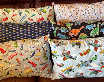 May include: A collection of rectangular throw pillows with various patterns. Designs include cars, trucks, airplanes, dinosaurs, and boats in a variety of colors. The pillows are arranged on a brown surface.