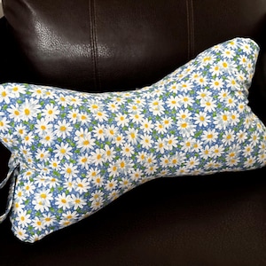 May include: A bone-shaped pillow with a blue background and a pattern of white daisies with yellow centers. The pillow has a loop on one end. The pillow is on a dark brown leather chair.