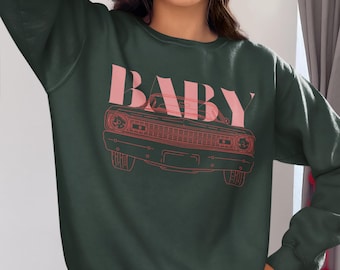 Könnte beinhalten: Dunkelgrünes Sweatshirt mit dem Wort "BABY" in rosa Blockbuchstaben über einer rosa Auto-Umrisszeichnung. Das Sweatshirt hat einen Rundhalsausschnitt und ist aus weichem Material.