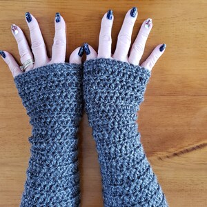 May include: A pair of gray crochet arm warmers. The handmade fingerless gloves are displayed on a wooden surface. The arm warmers are a solid gray color and have a textured, open weave design.