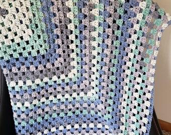 Puede incluir: Una manta de ganchillo en tonos grises, azules y verdes. La manta está hecha con un patrón de granny square y tiene un tacto suave y texturizado.