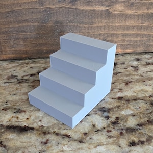 May include: A light gray, four-tiered staircase display stand with a smooth, matte finish. The structure features four distinct levels, each with a flat, rectangular surface, arranged in a stepped configuration to provide elevation for small items.