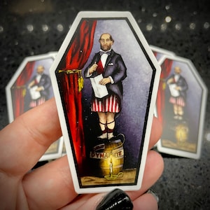 May include: A sticker featuring a man in a blue suit and red, white, and blue striped pants standing on a barrel labeled "Dynamite". The man is holding a scroll and is standing in front of a red curtain.