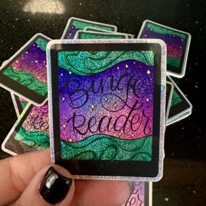 May include: A holographic sticker with a green, purple, and pink galaxy background. The sticker says "Binge Reader" in a white cursive font.