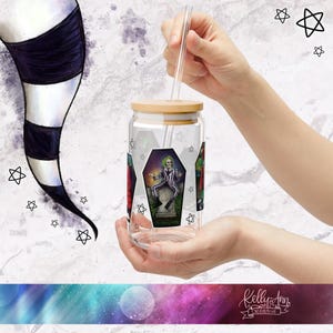 May include: A clear glass tumbler with a bamboo lid and a straw. The tumbler has a graphic of a character from the movie "Beetlejuice" sitting on a tombstone. The tumbler is being held by a hand.