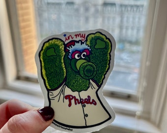 May include: A sticker featuring the Philadelphia Phillies mascot, a green furry creature with a baseball jersey that says "Pheeels". The sticker has the text "in my" above the mascot's head. The sticker is held up in front of a window.