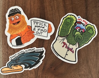 May include: Four stickers on a wooden surface. One features a cartoon character with orange fur and the text "PHUCK around & FIND OUT." Another shows a green mascot with "in my" above its head and "Pheeels" on its jersey. The third sticker is an eagle with the text "EMOTIONAL SUPPORT HOAGIE."