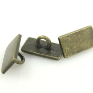 Puede incluir: Tres botones metálicos de color dorado antiguo con un bucle en la parte superior. Los botones son de forma rectangular.