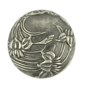 May include: A silver-toned metal button with a floral design. The button features a circular shape with a raised design of stylised flowers.