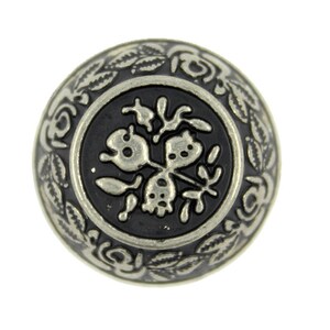 Puede incluir: Un botón plateado con un centro negro que presenta un diseño floral. El botón tiene un borde elevado y adornado con hojas y flores.