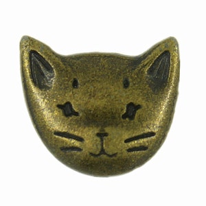 May include: A gold-toned metal button shaped like a cat's face. The button has a black outline and features a simple design of eyes, a nose, and whiskers.