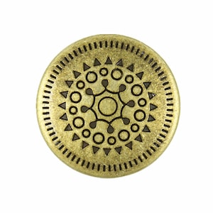 May include: A gold-toned metal button with a decorative design featuring a circle with a geometric pattern inside. The button has a textured surface and a raised edge.