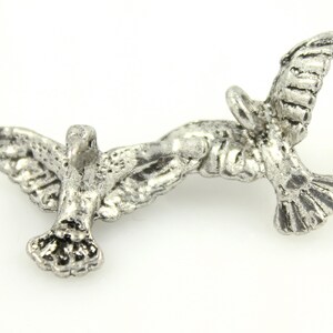 May include: Two silver doves in flight, facing opposite directions, with one dove's wings outstretched and the other dove's wings partially folded. The doves are joined together at the tail.